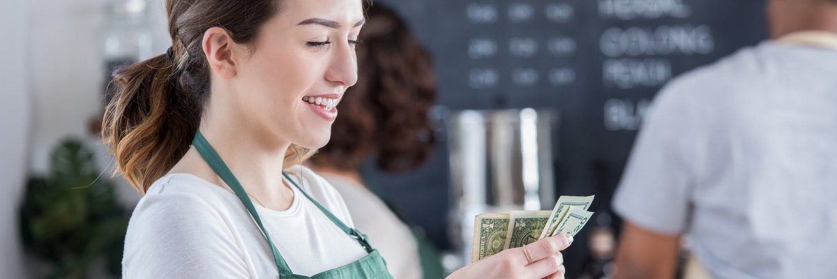 A smiling young barista counts cash in coffee shop.