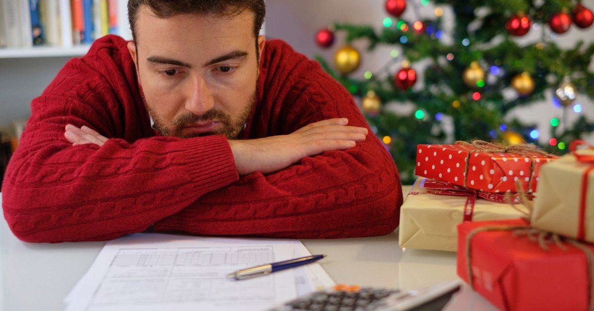 Man in festive sweater morosely slumped over desk looking at statements and calculator against Christmas tree backdrop.