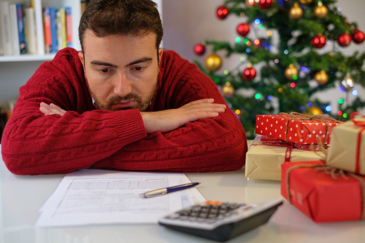 Man in festive sweater morosely slumped over desk looking at statements and calculator against Christmas tree backdrop.