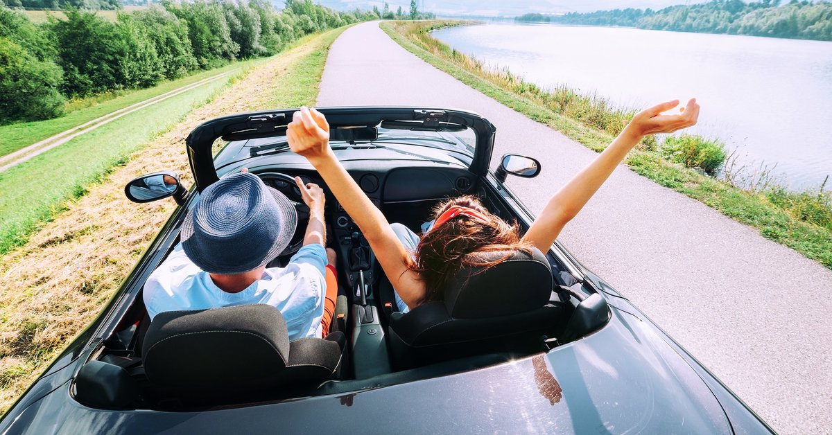 young man and woman riding in a convertible next to a lake on a sunny day.