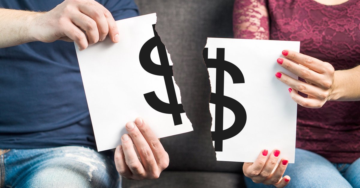 A couple sitting on a couch ripping apart a piece of paper with a big black dollar sign printed on it.