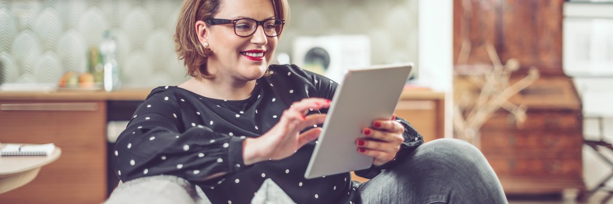Woman in glasses sitting on a couch smiling while using a tablet.