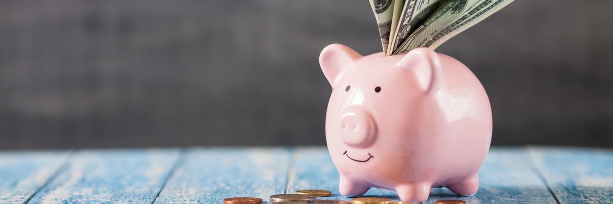 A piggy bank with folded bills sticking out the top and coins scattered around its feet.