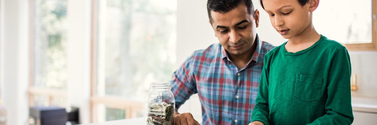 A parent and child count dollar bills and coins from a jar in their kitchen.