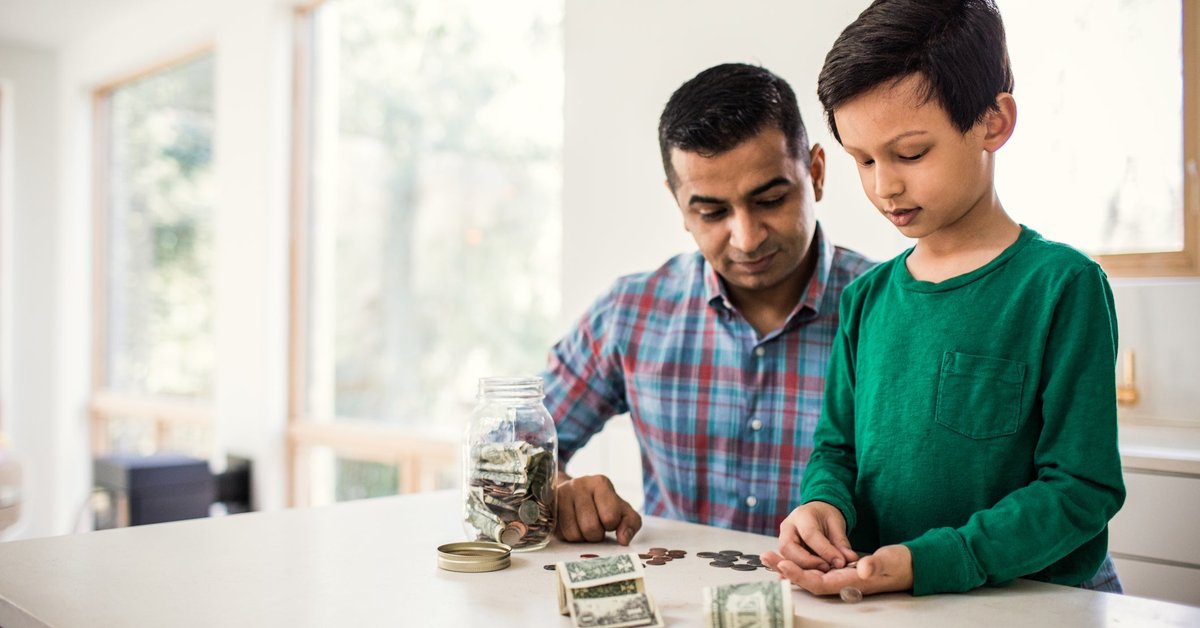 A parent and child count dollar bills and coins from a jar in their kitchen.