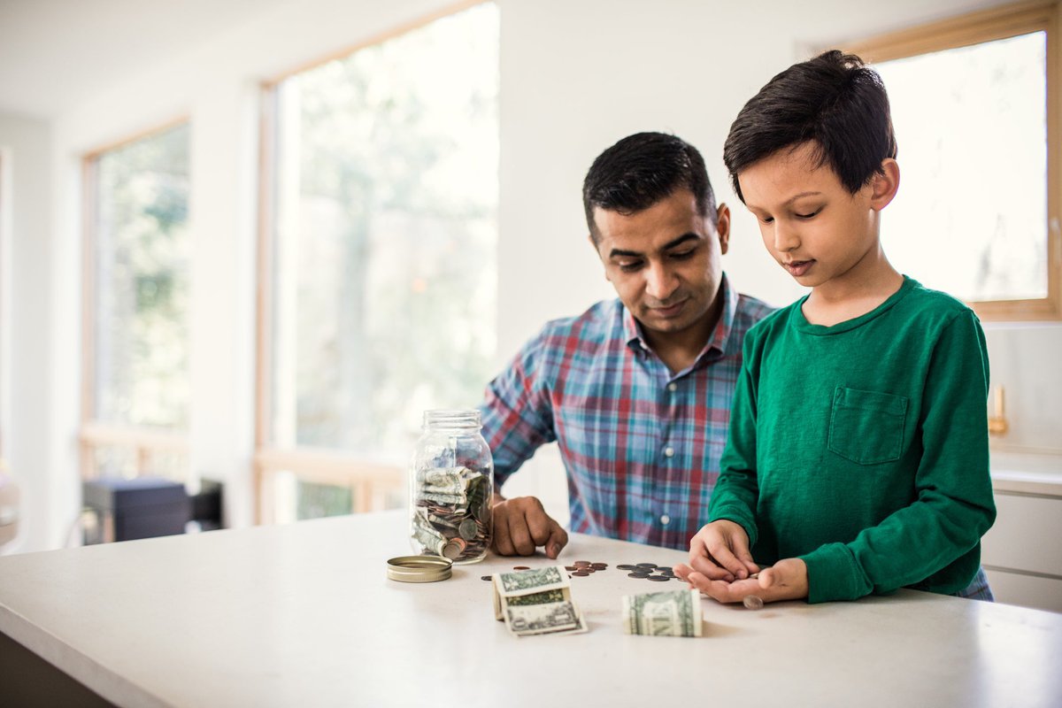 A parent and child count dollar bills and coins from a jar in their kitchen.