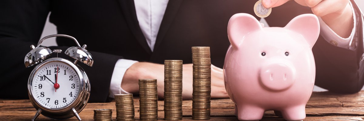 Person in suit puts coin into piggy bank next to stacks of coins and an alarm clock.