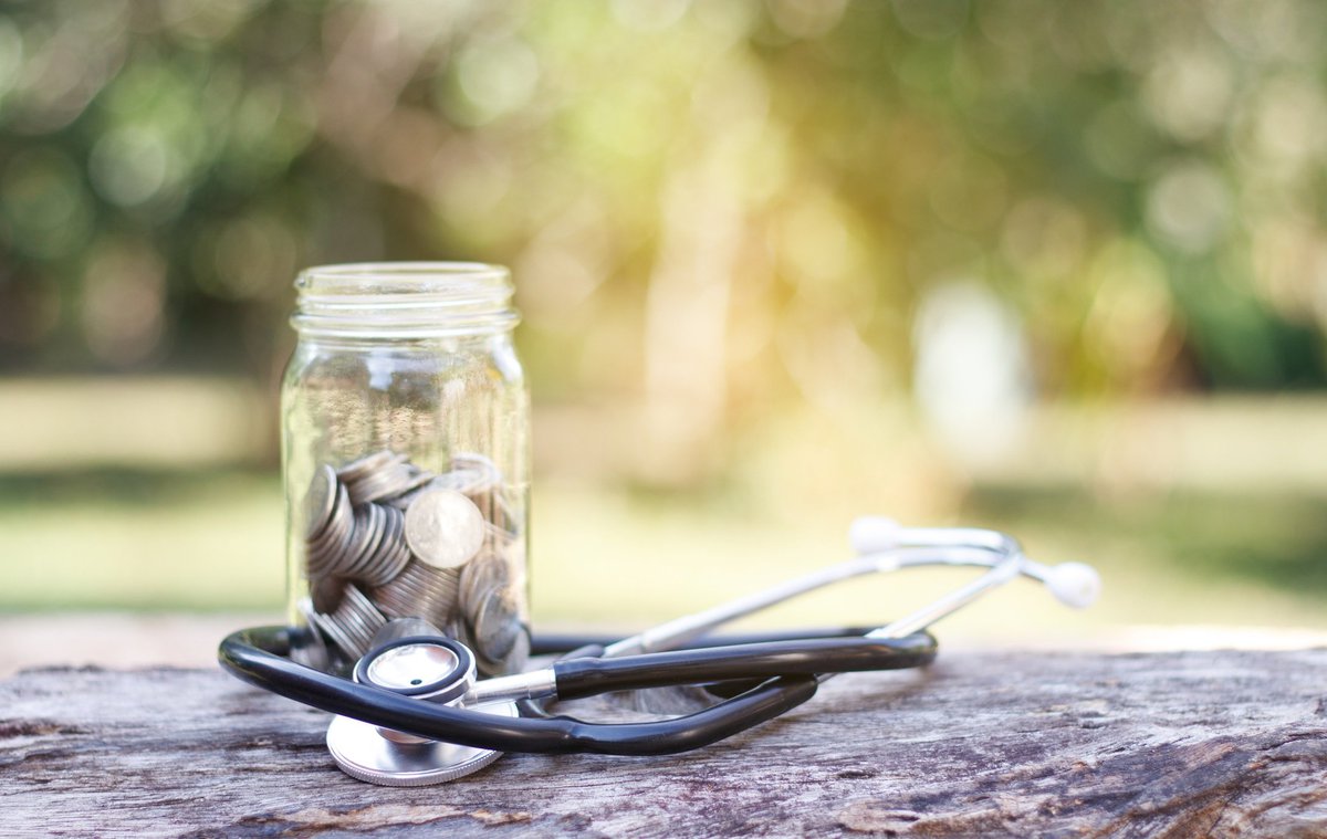 A jar full of coins sits with a stethoscope on top of a log outdoors.