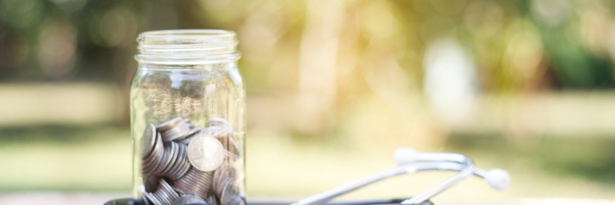 A jar full of coins sits with a stethoscope on top of a log outdoors.