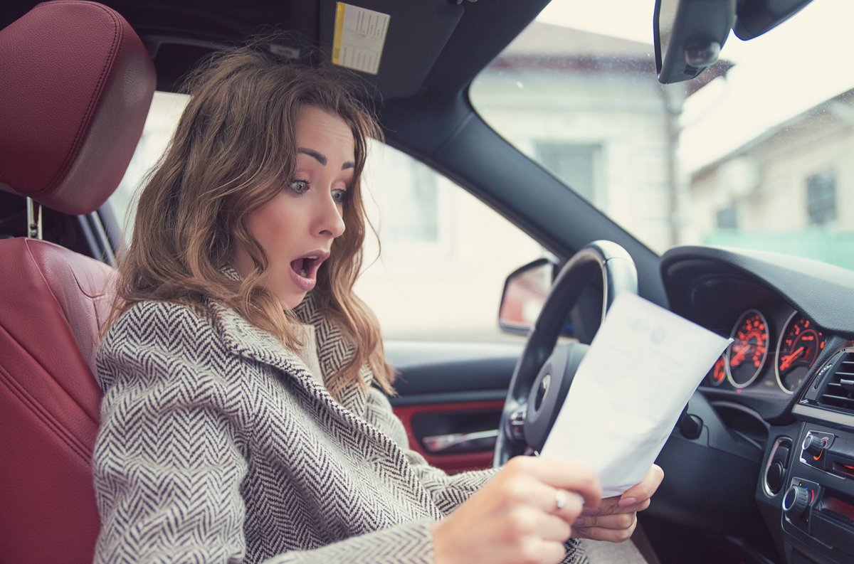 Woman sitting in car looking at piece of paper in shock.