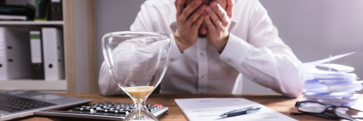 Person has there head in their hands at desk with an hourglass and calculator in the foreground.