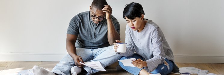 A couple sit on the floor in a room with no furniture surrounded by paperwork and bills.