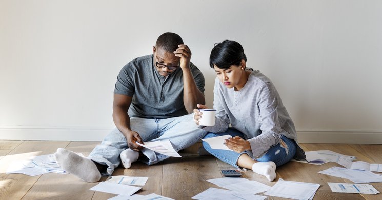 A couple sit on the floor in a room with no furniture surrounded by paperwork and bills.