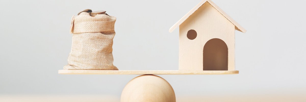 A toy house and a burlap bag of coins on opposite sides of a scale.