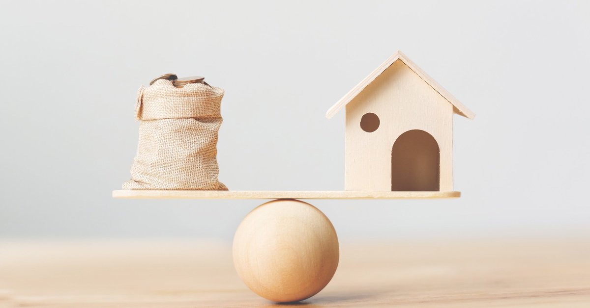 A toy house and a burlap bag of coins on opposite sides of a scale.
