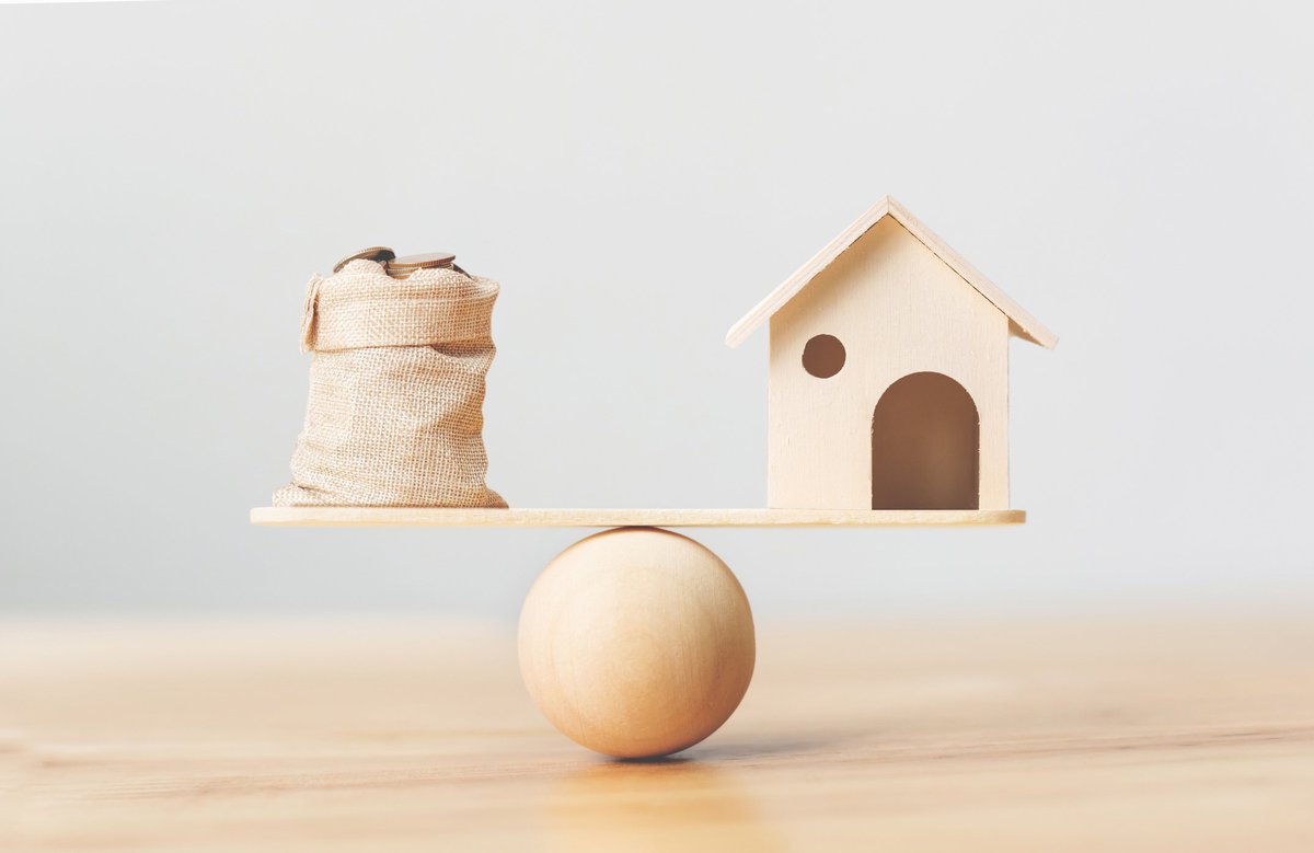 A toy house and a burlap bag of coins on opposite sides of a scale.