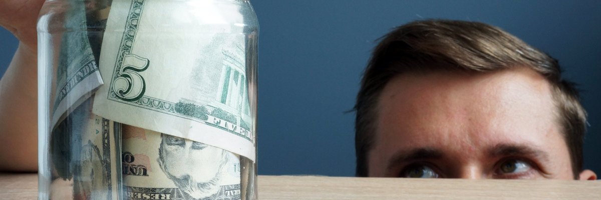 Half of man's face peering up over a counter as he sneaks a five-dollar bill out of a jar full of money.