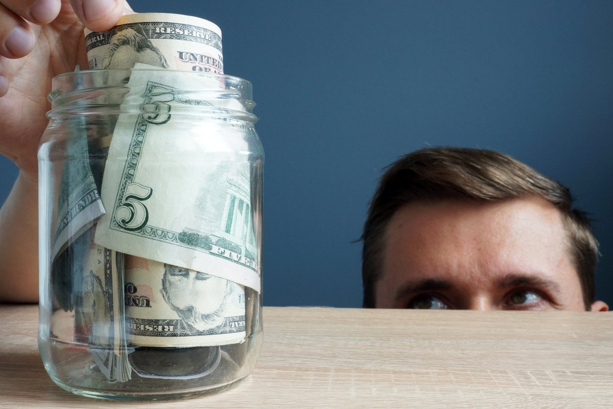 Half of man's face peering up over a counter as he sneaks a five-dollar bill out of a jar full of money.