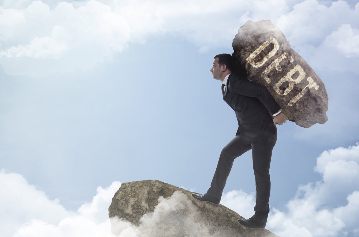 Man in suit carrying boulder labeled DEBT to edge of cloudy precipice.