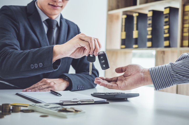 Man in suit reaching across desk to drop car keys into other person's hand.