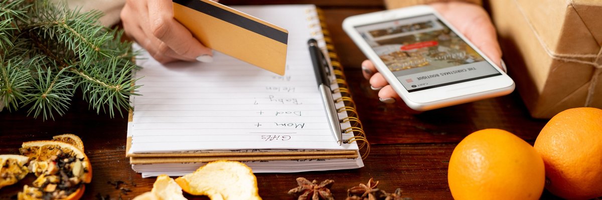 A woman writing a gift list while holding a credit card and phone with oranges and spices on the table in front of her.