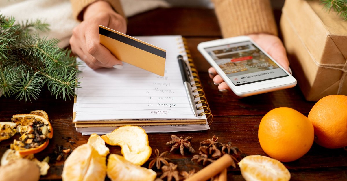 A woman writing a gift list while holding a credit card and phone with oranges and spices on the table in front of her.