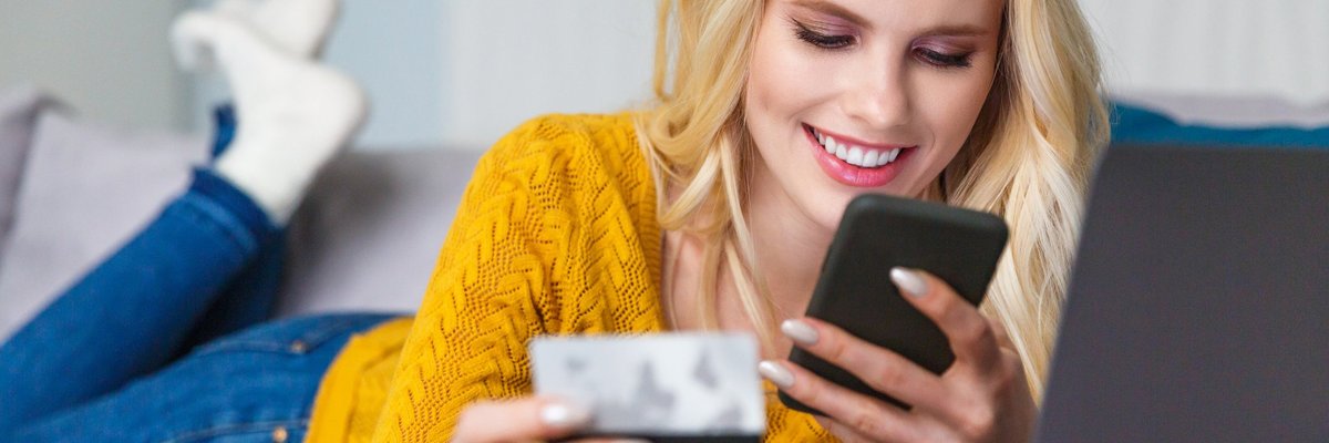Smiling woman lying on her couch, using her cell phone, and holding her credit card.