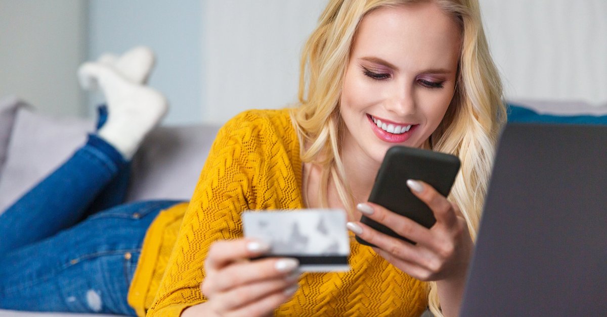 Smiling woman lying on her couch, using her cell phone, and holding her credit card.