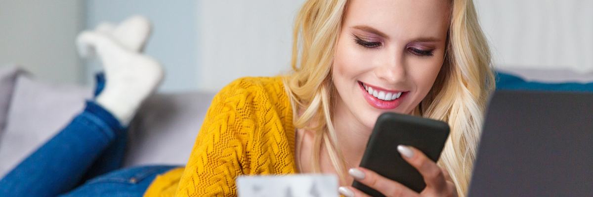 Smiling woman lying on her couch, using her cell phone, and holding her credit card.