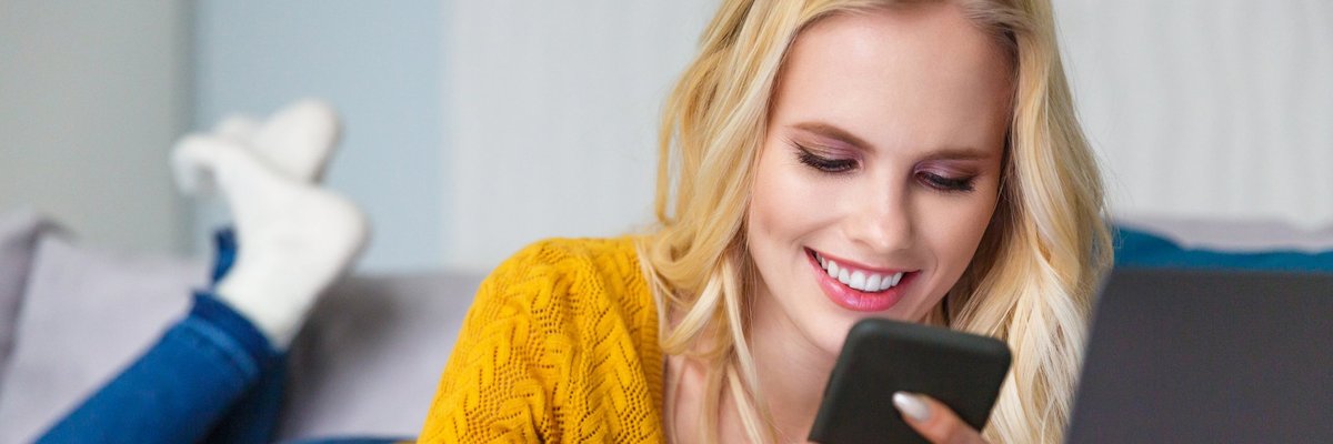 Smiling woman lying on her couch, using her cell phone, and holding her credit card.