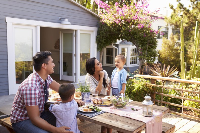 Two parents with their young children having lunch at a picnic table on their back deck.