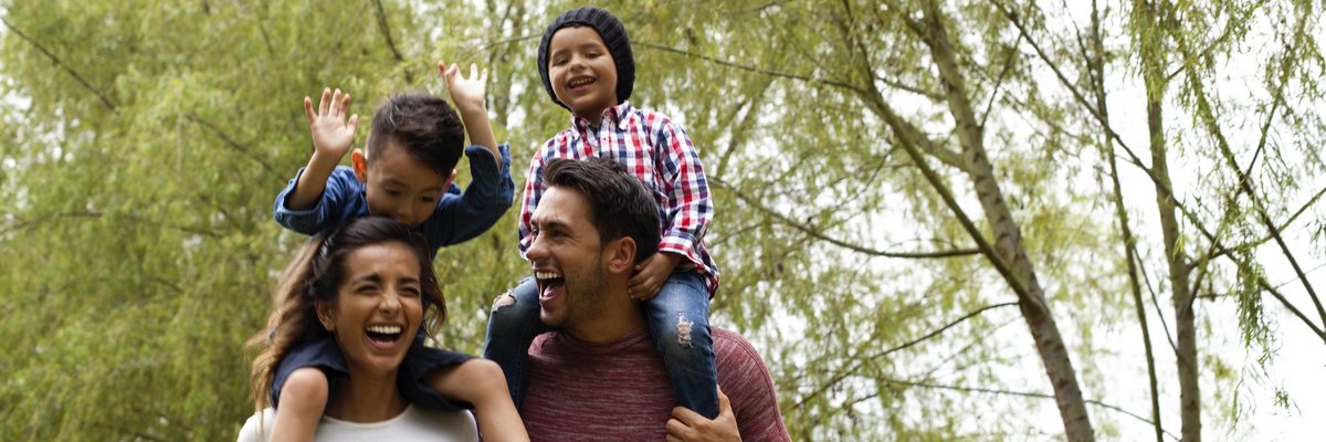 A happy family walking through a park with the young kids on the parents' shoulders.