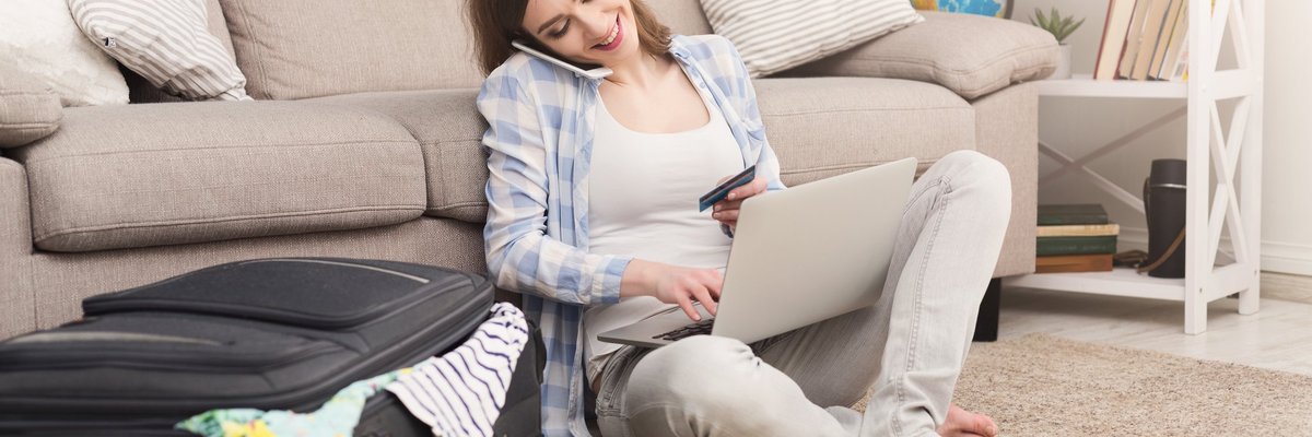 Woman sitting on floor beside suitcase talking on her cell phone and using a laptop.