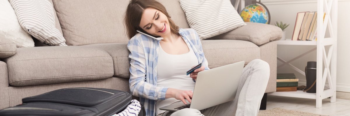 Woman sitting on floor beside suitcase talking on her cell phone and using a laptop.