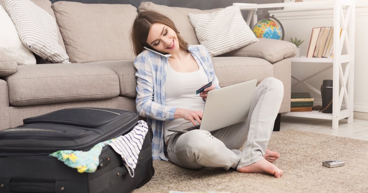 Woman sitting on floor beside suitcase talking on her cell phone and using a laptop.