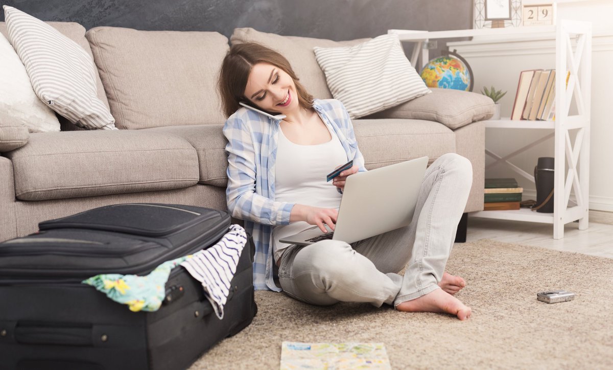 Woman sitting on floor beside suitcase talking on her cell phone and using a laptop.