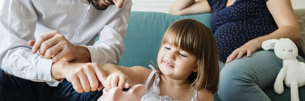 Happy Father Showing His Daughter How To Put Coins In Piggy Bank While Mom Watches