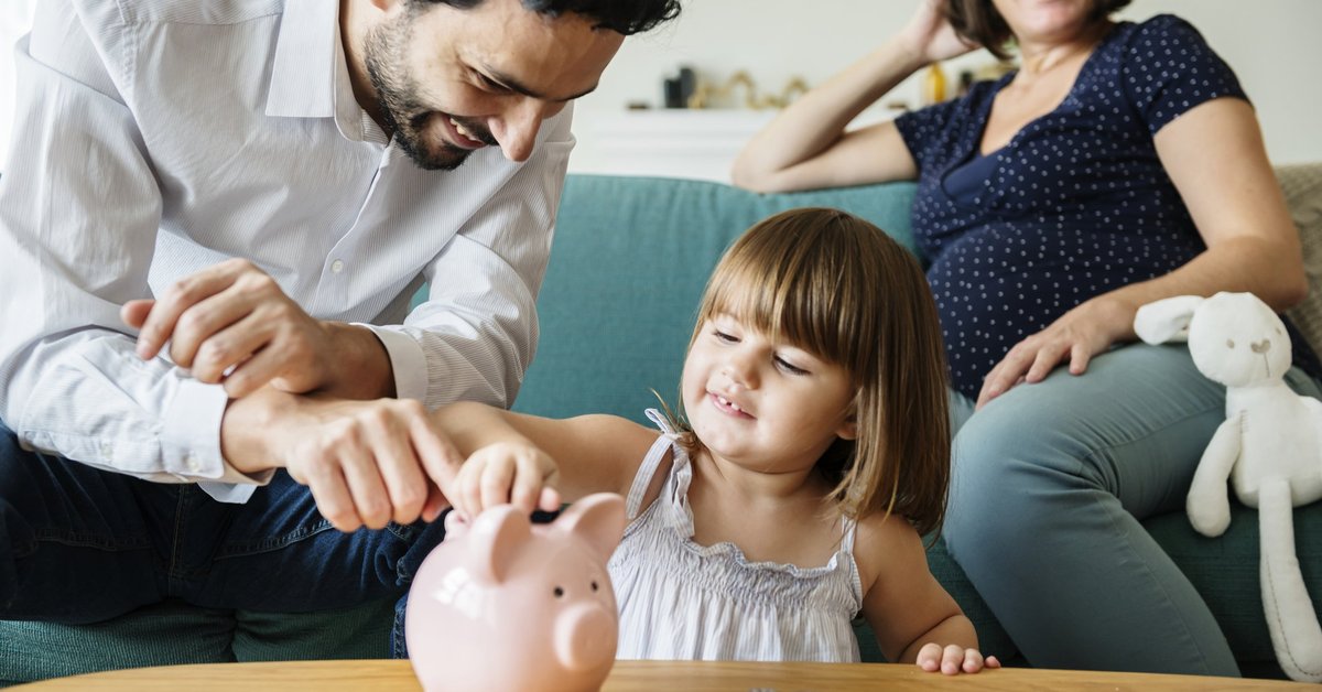 Happy Father Showing His Daughter How To Put Coins In Piggy Bank While Mom Watches