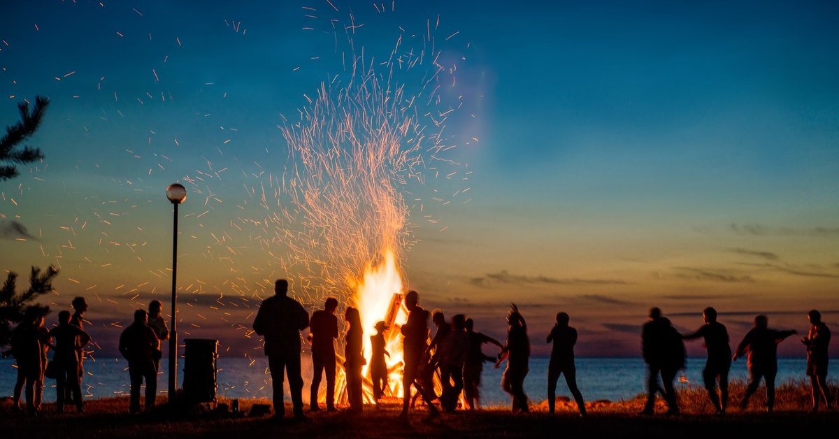 Large bonfire on a beach with a crowd gathered around.