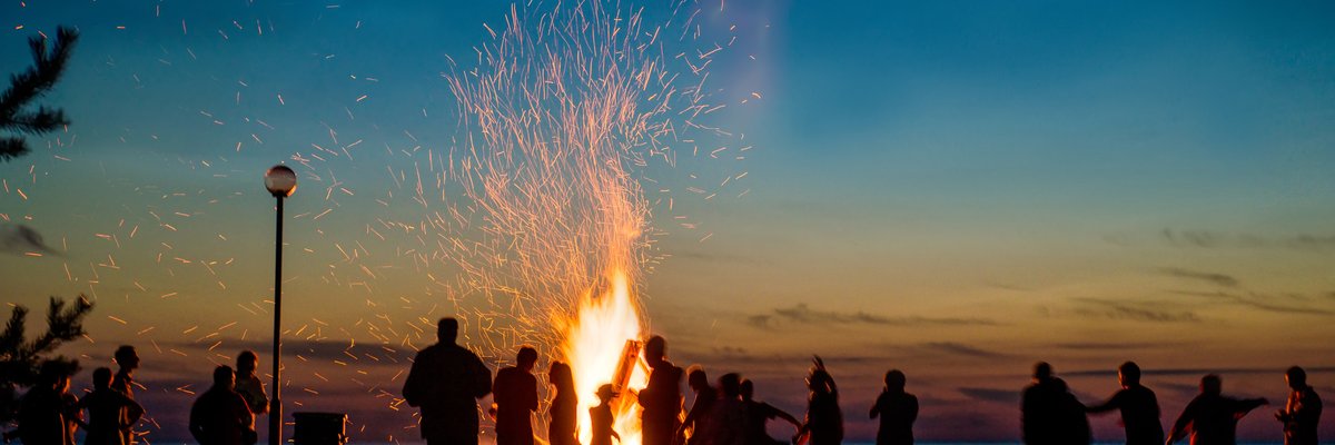 Large bonfire on a beach with a crowd gathered around.