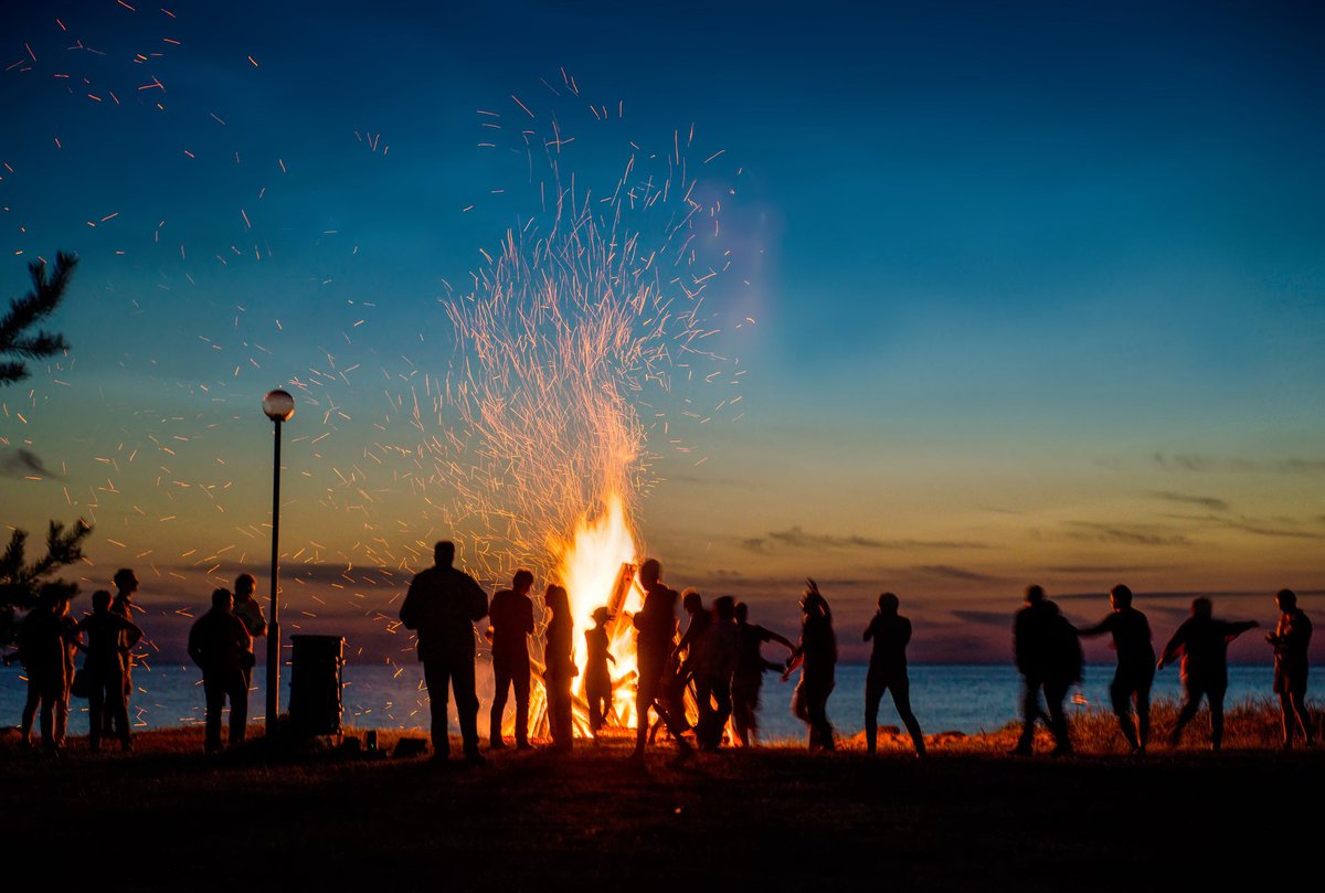 Large bonfire on a beach with a crowd gathered around.