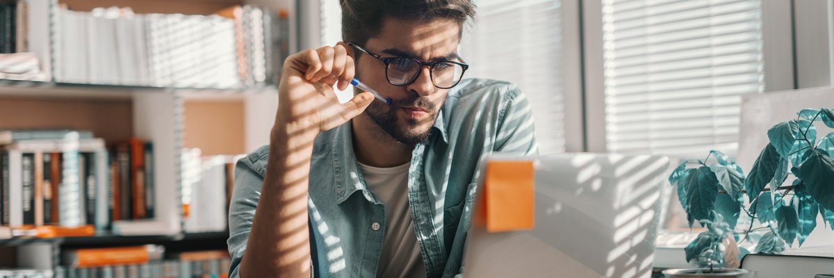A young adult sitting at a sunny desk and looking at their laptop.