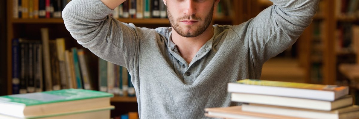 A stressed male college student with a stack of books in the library.
