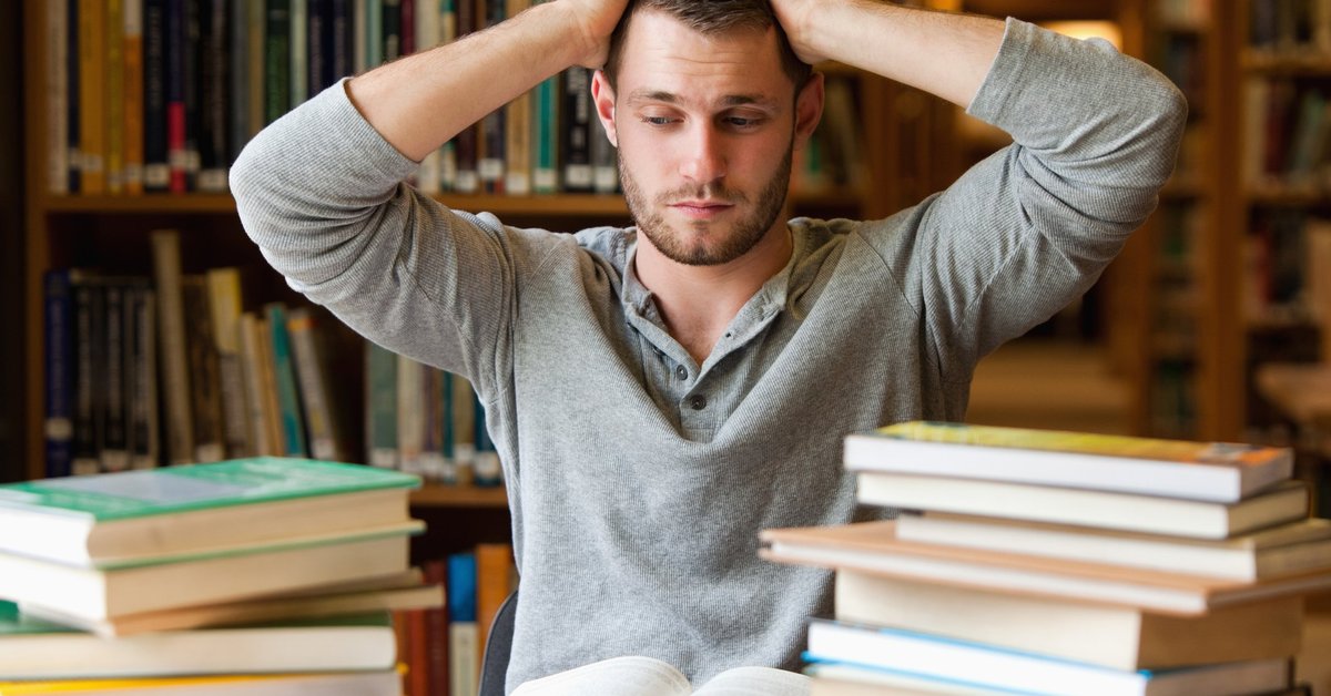 A stressed male college student with a stack of books in the library.
