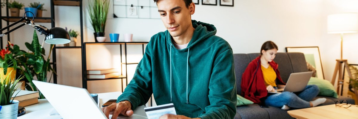 At home, a man sits at a table and is using his computer while holding his credit card.