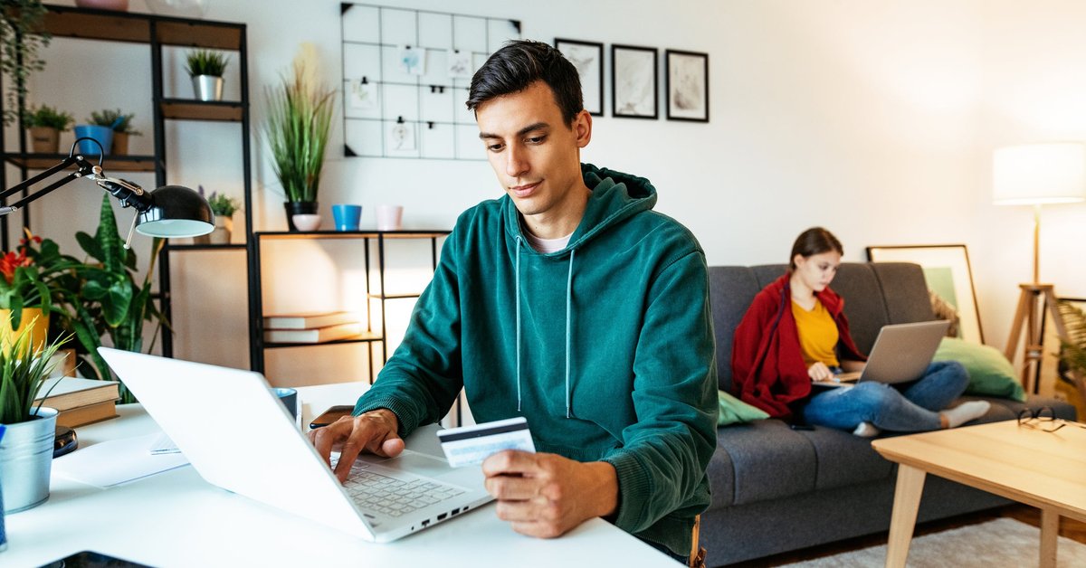 At home, a man sits at a table and is using his computer while holding his credit card.