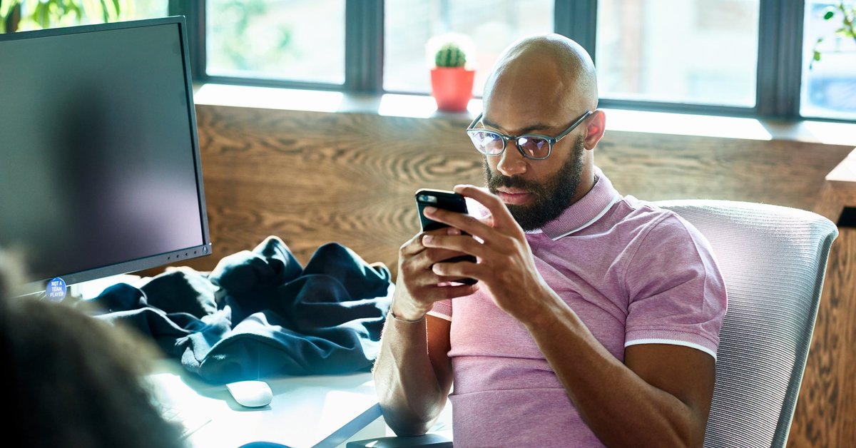 A person sitting at an office desk and looking at a cell phone with a contemplative expression.