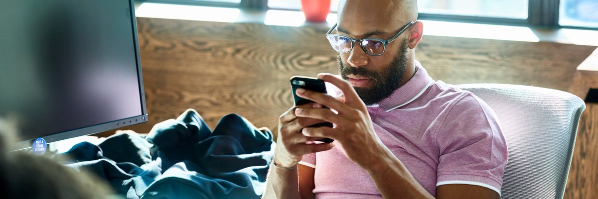 A person sitting at an office desk and looking at a cell phone with a contemplative expression.