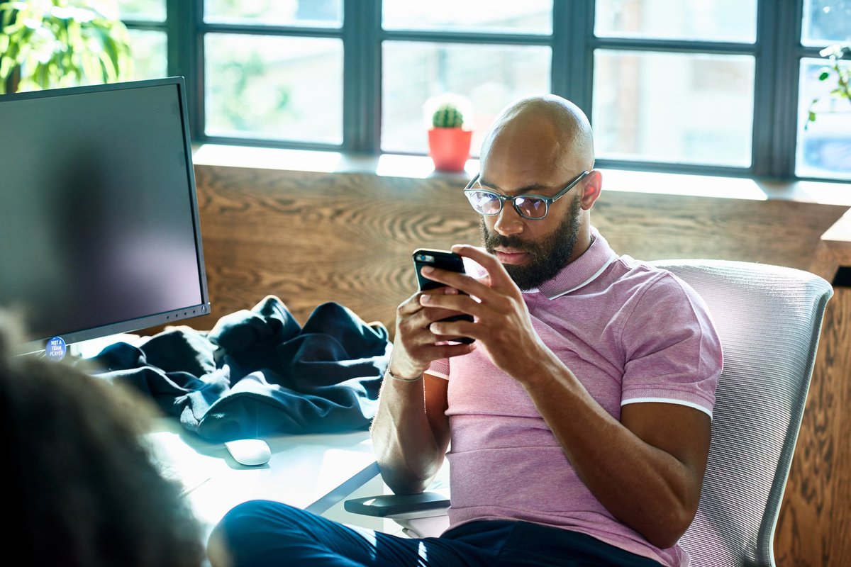 A person sitting at an office desk and looking at a cell phone with a contemplative expression.