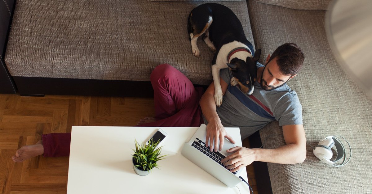 A man sitting on the floor in front of his couch with his dog looking over his shoulder at his open laptop.